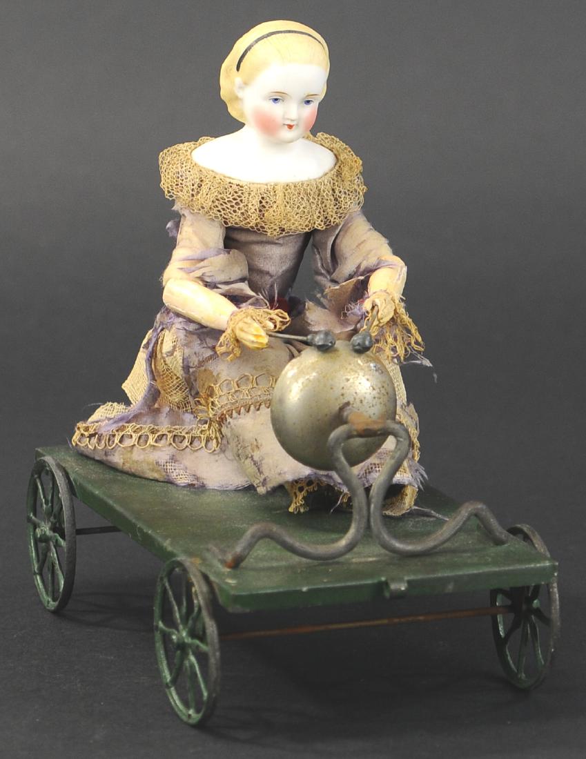 BELL CHIME PULL TOY/DOLL: 201. BELL CHIME PULL TOY/DOLL | Sweet early 1870s German Parian head doll with wooden arms, seated to a painted green platform with arms outstretched and mallets in hand, as toy is pulled along, the t
