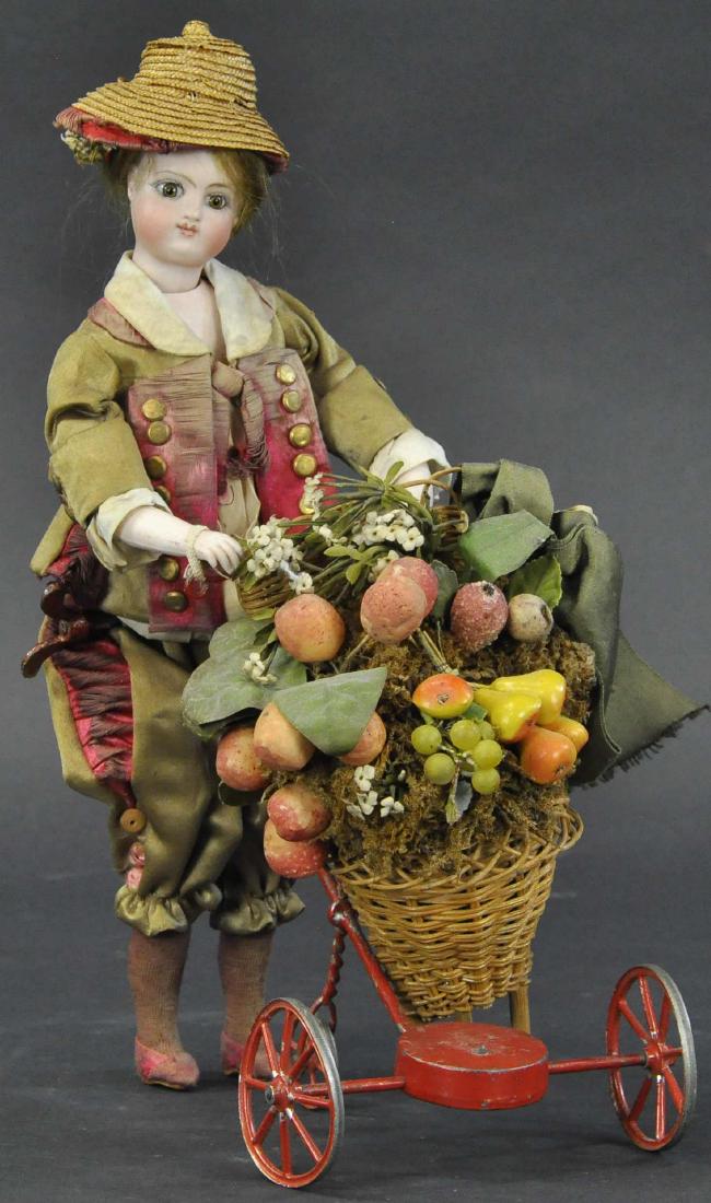 FRUIT SELLER MECHANICAL DOLL/TOY: 252. FRUIT SELLER MECHANICAL DOLL/TOY | Impressive all original standing/walking figure with bisque head and early "Jules Steiner" characteristics, wearing fancy silk costume and hat pushing a tall ta