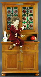 "LE BUFFET MAGIQUE" MUSICAL AUTOMATON: 254. "LE BUFFET MAGIQUE" MUSICAL AUTOMATON | Elaborate unconventional automaton of a boy with mache head seated on the mid level shelf of a wooden kitchen cabinet, holding a mache "piece of bread" in