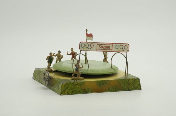 OLYMPIC STADIUM TOY: Doll, Germany, hand painted tin circular track on square base, features Olympic Marquee and five composition figures on individual supports, steam accessory flywheel on side. 11" x 3" without flag.