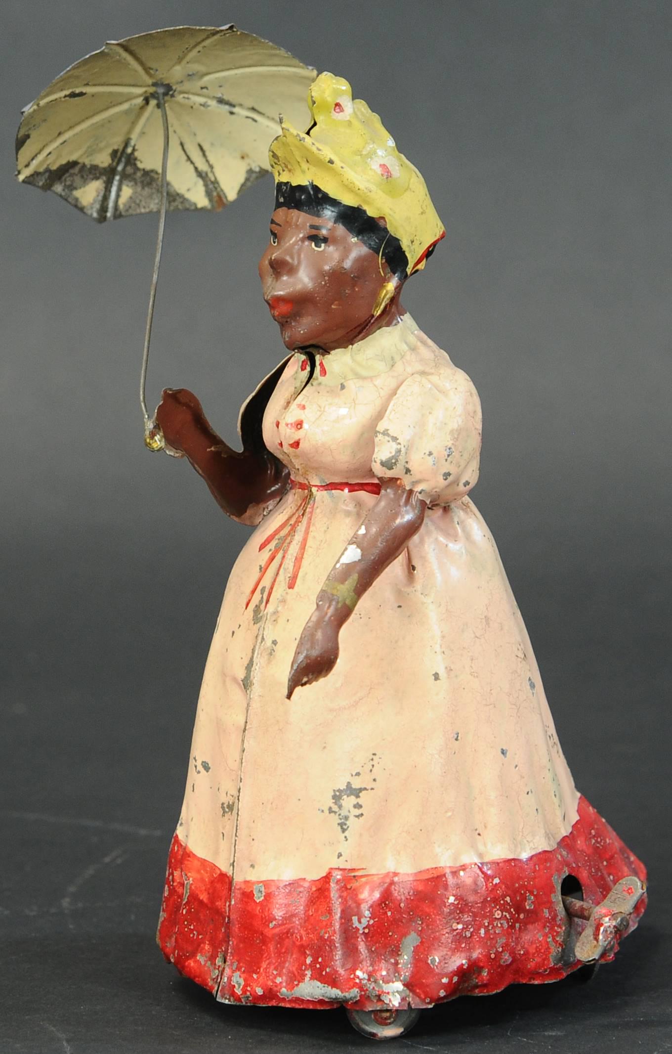 HANS EBERL WOMAN W/ UMBRELLA: 114. HANS EBERL WOMAN W/ UMBRELLA | Nice German toy with a large shelf presence, hand painted tin, key wound with a bellows emitting squeak noises as she seemingly walks. | Provenance: Paul Cole Colle