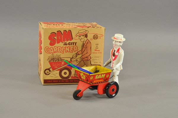SAM THE GARDNER WITH ORIGINAL BOX: Louis Marx, lithographed tin open wheel barrow pushed by plastic figure of Sam the Gardner, complete with three plastic gardening tools, key wind operated, walking motions. 7 1/2" l.