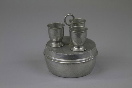 HUNTLEY & PALMERS "EGGSTAND" BISCUIT TIN: England, c. 1910 Rare 'hammered aluminum' container featuring circular dish with lift off lid fitted with handle and pegs to accommodate three egg cups. 6 ½" h.