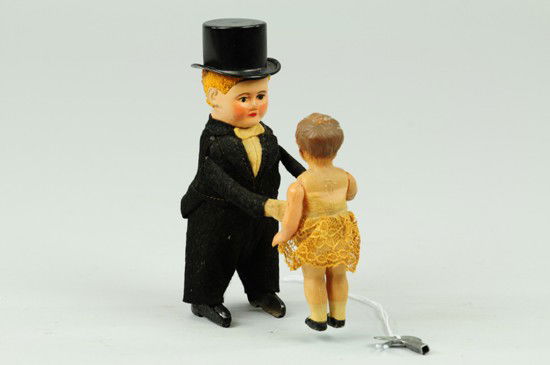 BOY IN TUX WITH GIRL: Schuco, Germany, clockwork driven, boy dressed in black tuxedo and tin top hat, white shirt and bow tie, celluloid girl with cream lace dress. 5" h.