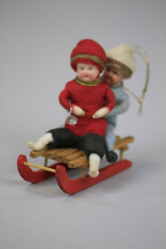 TWO PRESSED COTTON CHILDREN ON SLED: Germany, two pressed cotton children with celluloid faces, hats and necks trimmed in golden Dresden ribbons, dressed in very colorful red and blue sledding outfits, a horn in first child's hand, both 