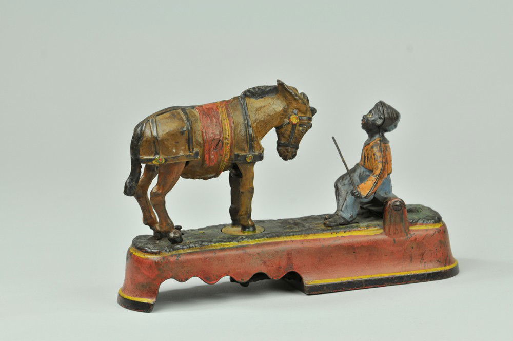 SPISE A MULE MECHANICAL BANK (BENCH): Orange shirt, J & E Stevens Co., designed by James H. Bowen, patented 4/27/1897. When the mule and boy are brought into position, a slight touch on the knob at the base causes the mule to turn around