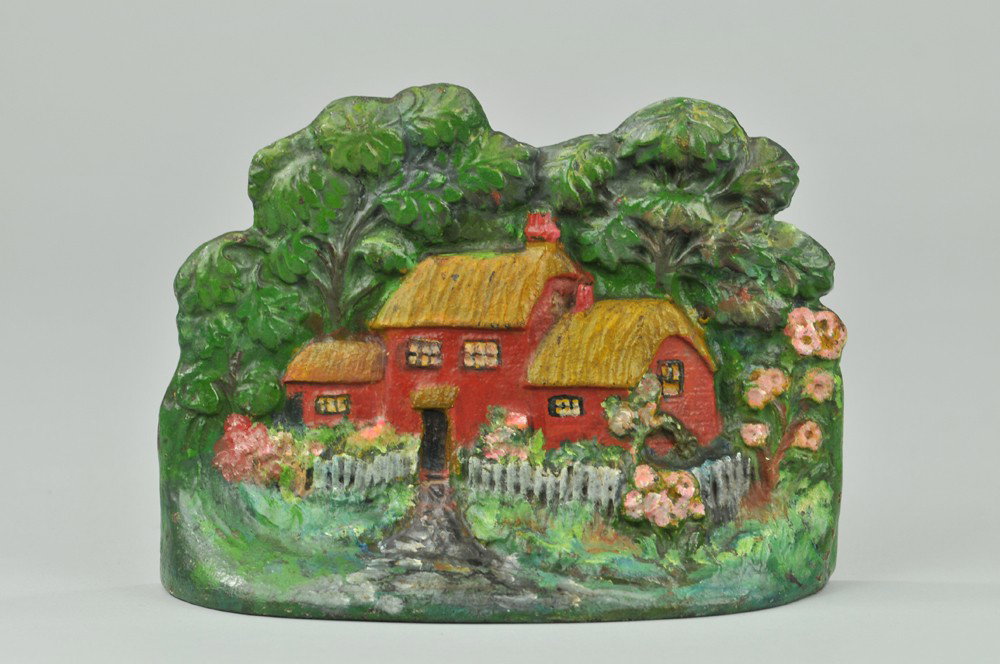 TWO-STORY THATCHED ROOF COTTAGE DOORSTOP: "Crenier Novelties, No. 8," inscribed on back. Fully forested scene of red house w/white picket fence, accented by pink flowers throughout. Cobblestone trail leading to house focal point. Very rare, h