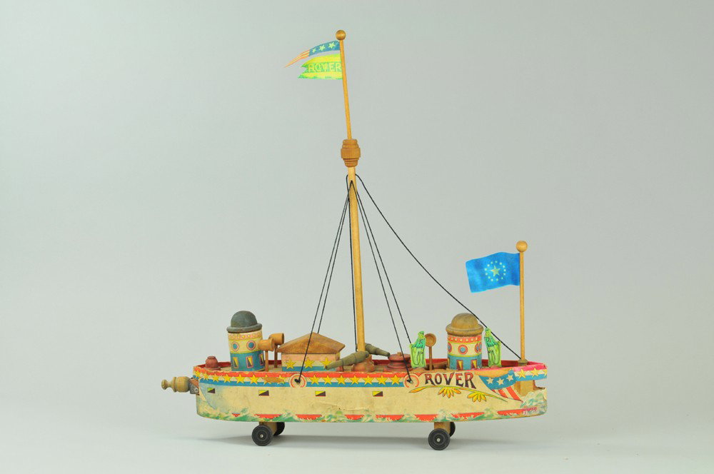 BLISS BATTLESHIP ROVER: Lithographed paper over cardboard hull, upper deck is quite detailed observation towers, center mast, guns aboard. 19" l. Laser copy repairs at hull and flags, overall (Fair Cond) 19" l.
