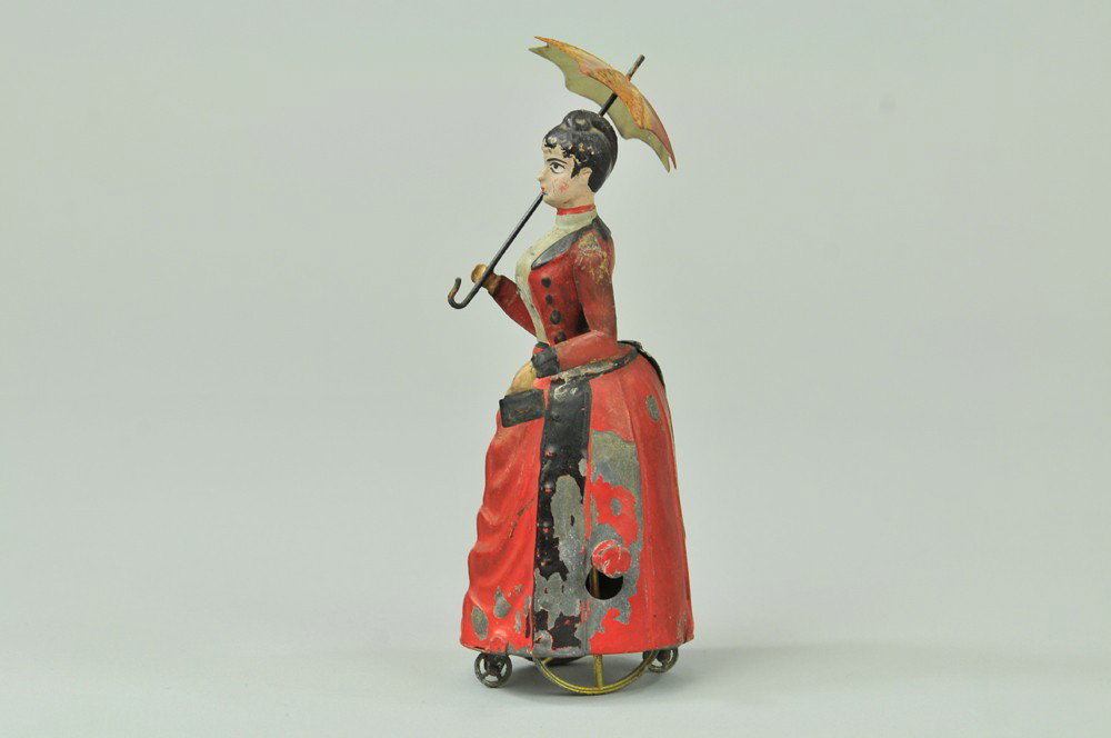 WOMAN WITH UMBRELLA: Attributed to French toy maker, hand painted tin, well detailed overall, features woman holding umbrella in hand, spoke wheel base and fly wheel appears on side. 4" h. Repainted. 4" h.