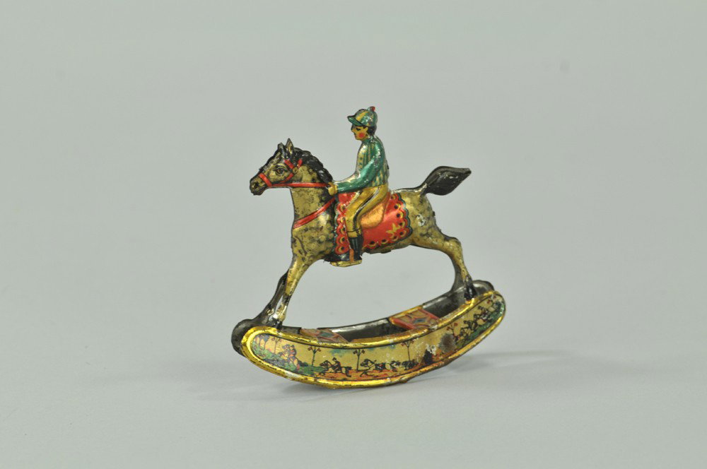 JOCKEY ON HORSE ROCKING PENNY TOY: Meier, Germany, lithographed tin, rocker base depicts steeple chase theme, seated jockey, very colorful. 3 3/4" l. Wear to rocker base, otherwise (Exc. Cond) 3 3/4" l.