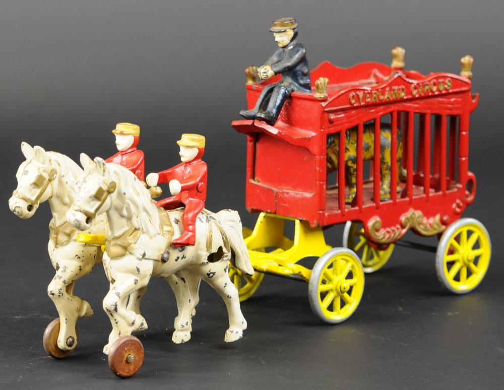 OVERLAND CIRCUS CAGE WAGON: Cast iron, enclosed cage wagon painted in red with gold trim, houses yellow lion, driver seated on rooftop, yellow spoke wheels, drawn by two white horses and mounted riders. 14" l.