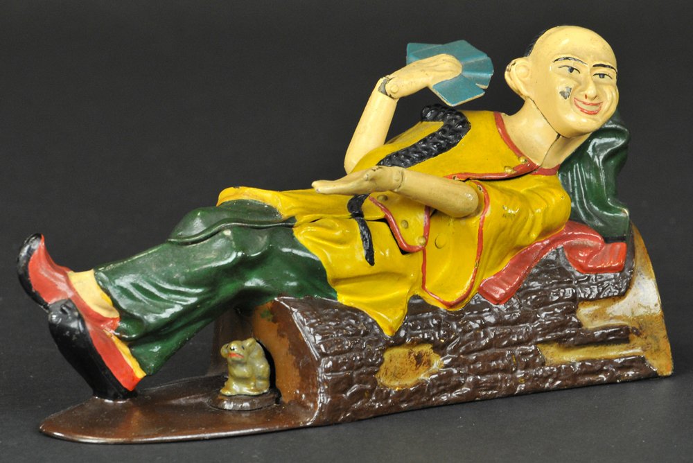 RECLINING CHINAMAN MECHANICAL BANK: J & E Stevens Co., designed by James H. Bowen, circa 1885. Place the coin in the opening. Press the lever, and the Chinaman will show his hand (four aces) and salute the depositor. As the coin falls i