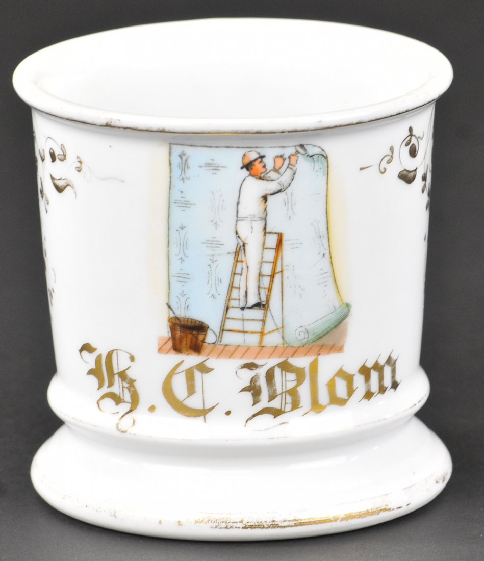 WALLPAPER HANGER OCCUPATIONAL SHAVING MUG: Number markings on base, features man standing on ladder hanging pastel colored wallpaper, gold gilt name reads, "H. C. Blom," floral décor painted on sides, gold loss to base rim. Ex. Bill Bertoia C