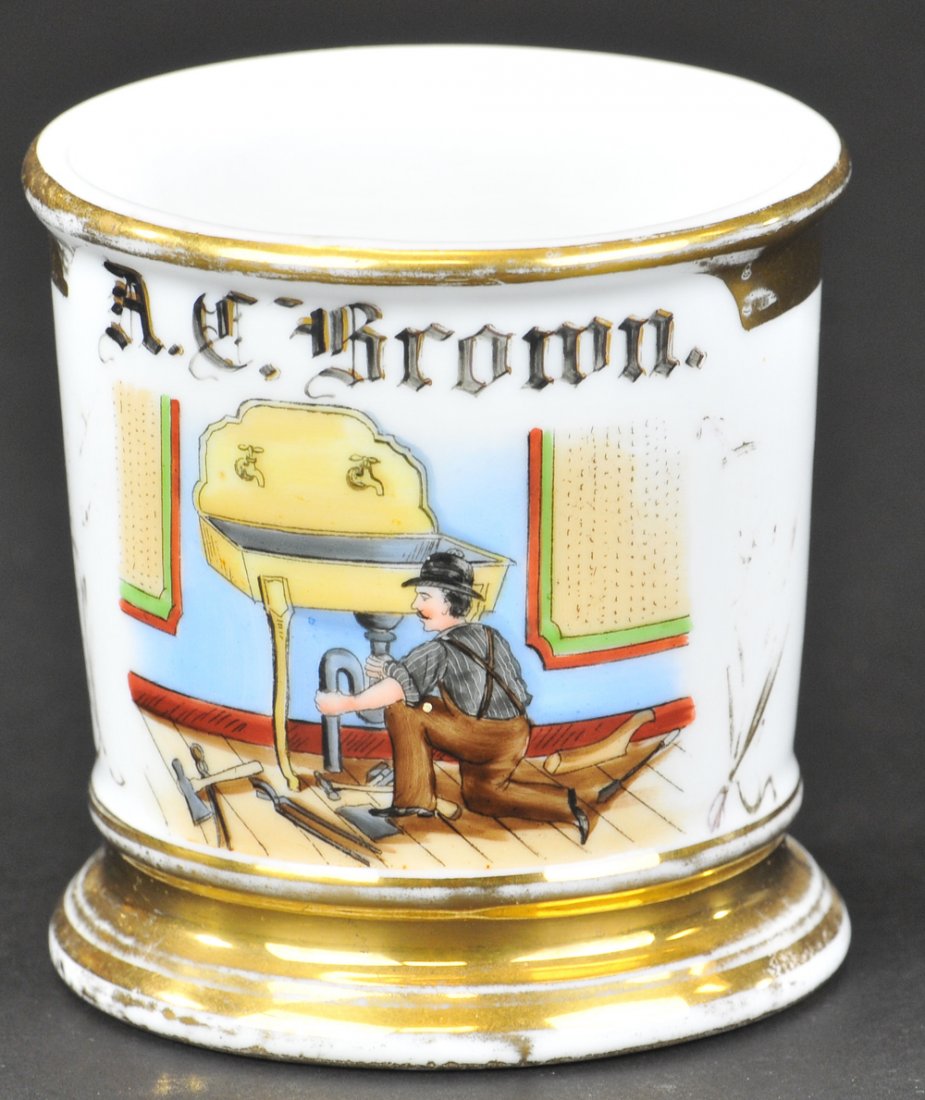 PLUMBER OCCUPATIONAL SHAVING MUG: Unmarked base, wonderful artistry illustration of a working plumber at sink, tools lie on floor, great colors, name reads "A. C. Brown," full gold base rim. Ex. Bill Bertoia Collection 3 5/8" h.