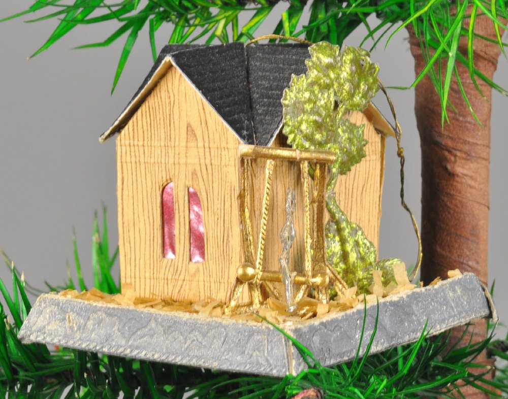 DRESDEN MAKER'S COTTAGE CHRISTMAS TREE ORNAMENT: Germany, a Dresden maker's forest cottage with tree and person on swing, red windows in wood grain siding. 2 1/2" l. Ex. Tom Fox Collection.