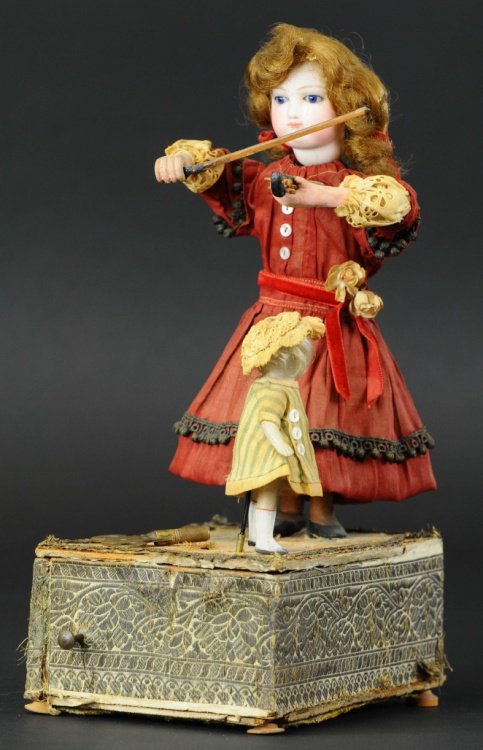 YOUNG GIRL PLAYING FIDDLE WITH TINY DANCER: c. 1895, attributed to French company Renou, bisque head girl holds wooden violin while tiny bisque jointed figure twirls around on square base as clockwork is activated. Ex Richard Pryor Collection.