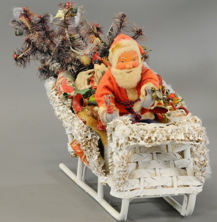 SANTA AND SLEIGH LOADED WITH TOYS: A composition Santa dressed in red top and blue pants sits in front a lichen trimmed wicker sleigh loaded with an impressive amount of Christmas toys, topped off by decorated feather tree making for a