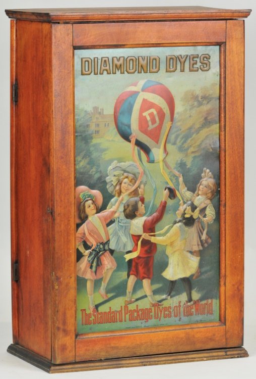 COUNTER TOP DIAMOND DYE CABINET: Embossed tin litho scene of children in brightly colored clothes dancing with a hot air balloon, the Standard Dyes of the World, in cherry wood cabinet. 24" h.