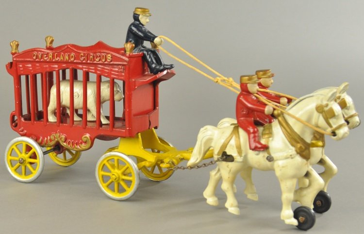 KENTON OVERLAND CIRCUS CAGE WAGON: Cast iron, enclosed cage wagon painted in red with gold trim, houses white bear, driver seated on rooftop, yellow spoke wheels, drawn by two white horses and mounted riders. 14" l.