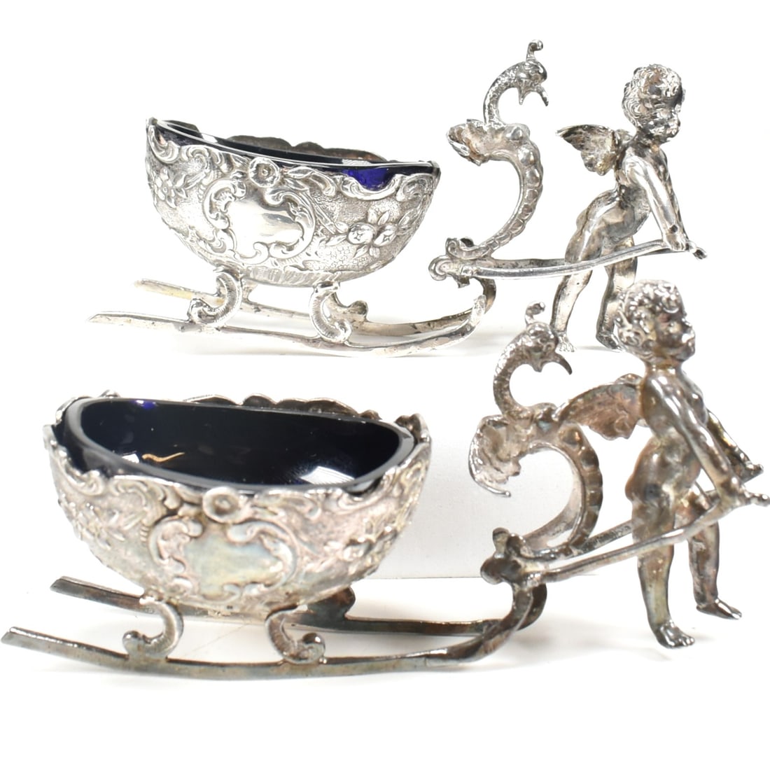 925 SILVER PAIR OF BLUE GLASS LINED CHERUB SLEIGH TABLE SALTS TBV: 925 silver pair of blue glass lined cherub sleigh table salts. Each with blue glass lined salt sleighs pulled by winged cherub - putti. Stamped with sponsors mark BM, 925 with oak leaf assay mark and