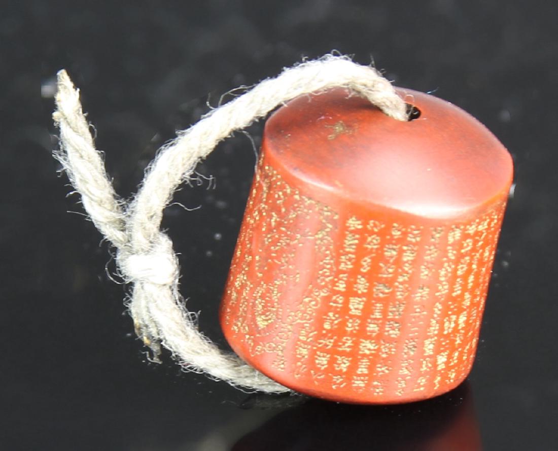 Chinese Red Sand Bead: China, 19th C., red sand bead, in cylindrical form, with gilt and incised Buddhist inscriptions across the surface, in a brick red hue, with rope. Height 3/4 in.