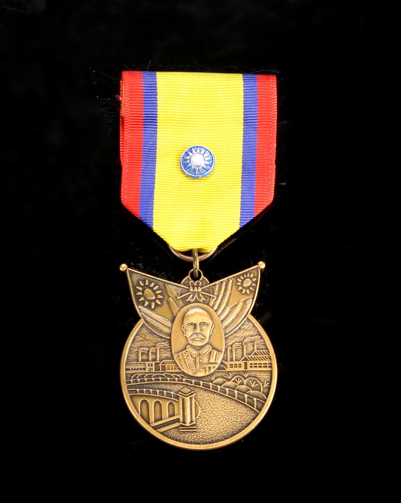 Chinese Military Badge: China, ROC Period, military badge, circular in form, with winged top, image of a figure in background factory scene, red, blue and yellow ribbon. Diameter 1 3/4 in.