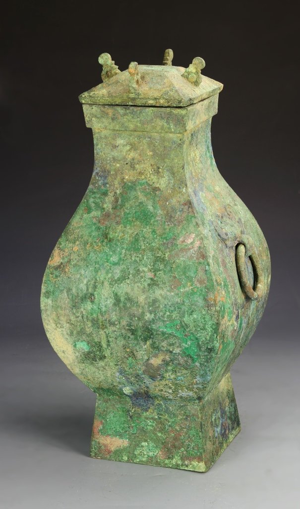 Chinese Han Dynasty Bronze Vase: China, Han Dynasty, bronze vase in a squared footed form, with rings emerging from masks at the side, cover with four projecting decorative elements, heavy patina overall. Height 18 in.