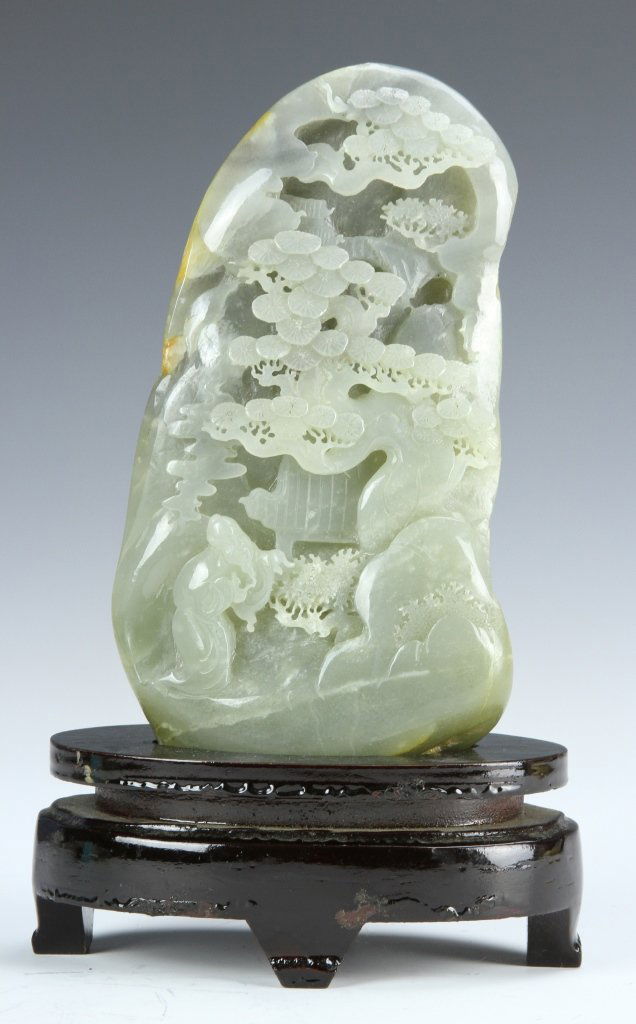 Chinese Hetian Jade Mountain: China, Hetian jade piece in the form of a mountain, carved to show a bearded figure, standing among tree branches, presented on a two tiered wooden stand. Height: 6 3/4 in.