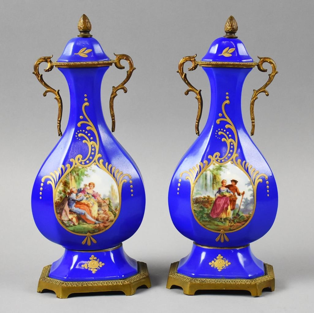 Pair French M. Sèvres Porcelain & Gilt Bronze Covered Urns: These are a beautiful pair of Sevres style cobalt painted covered vases. Lidded and are hand painted, each featuring a courting scenes surrounded by hand painted scrolls, flowers and leaves. Acorn fin
