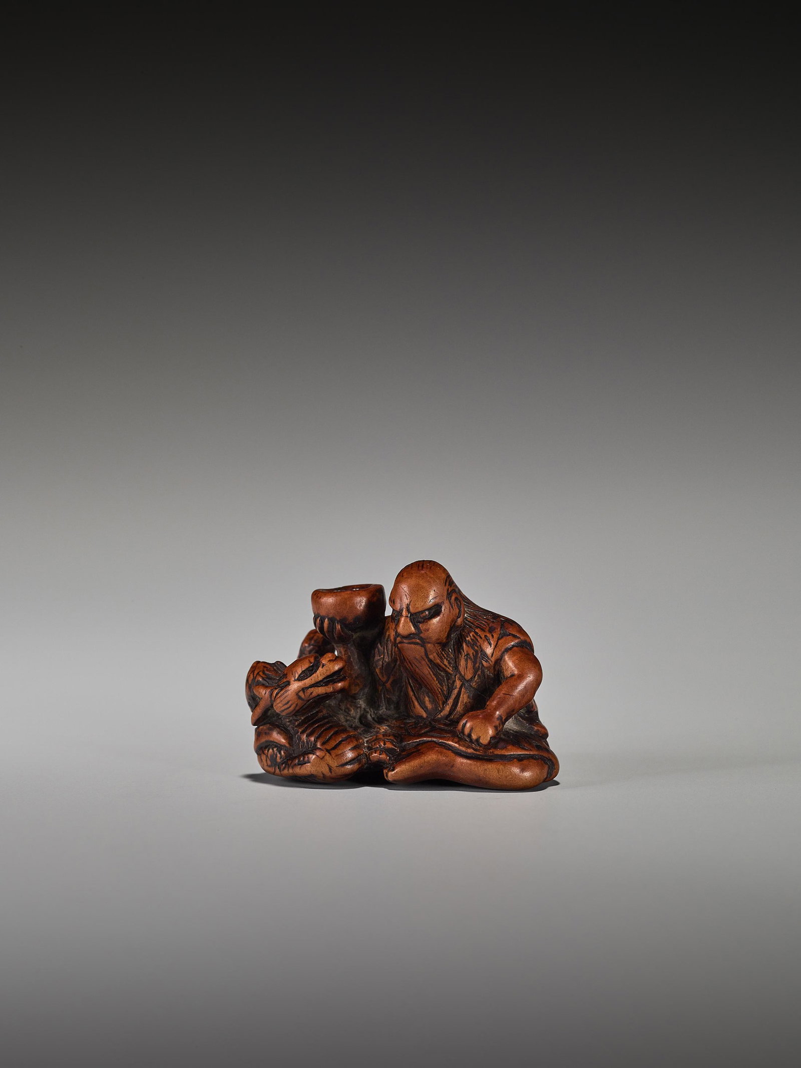 A RARE WOOD NETSUKE OF CHINNAN SENNIN: A RARE WOOD NETSUKE OF CHINNAN SENNIN Unsigned Japan, 19th century, Edo period (1615-1868) Chinnan Sennin is depicted seated holding his vessel, which he used to conjure the dragon lying next to him, 
