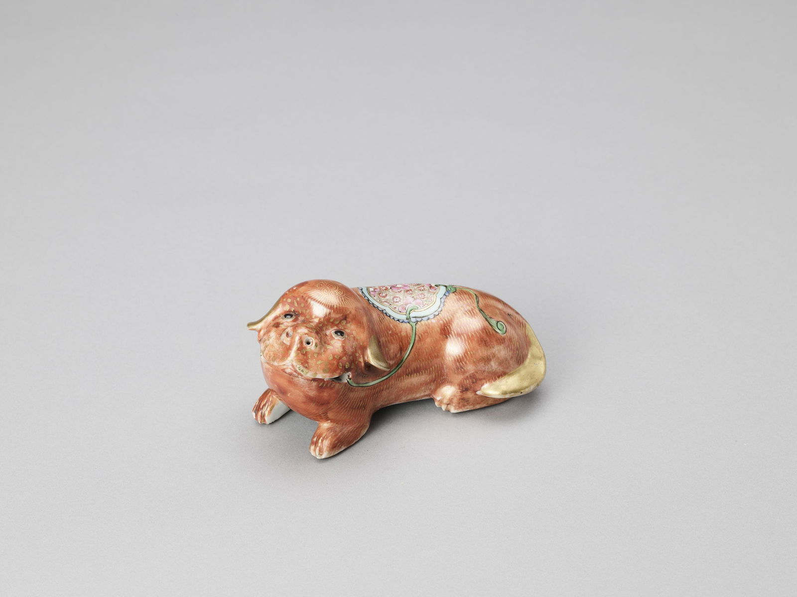 A FAMILLE ROSE PORCELAIN DOG, QING: A FAMILLE ROSE PORCELAIN DOG, QING China, earlier 19th century. The recumbent dog finely modelled and painted in iron red, gold and vivid enamels. Good detail work to saddle and fur. Condition: Small