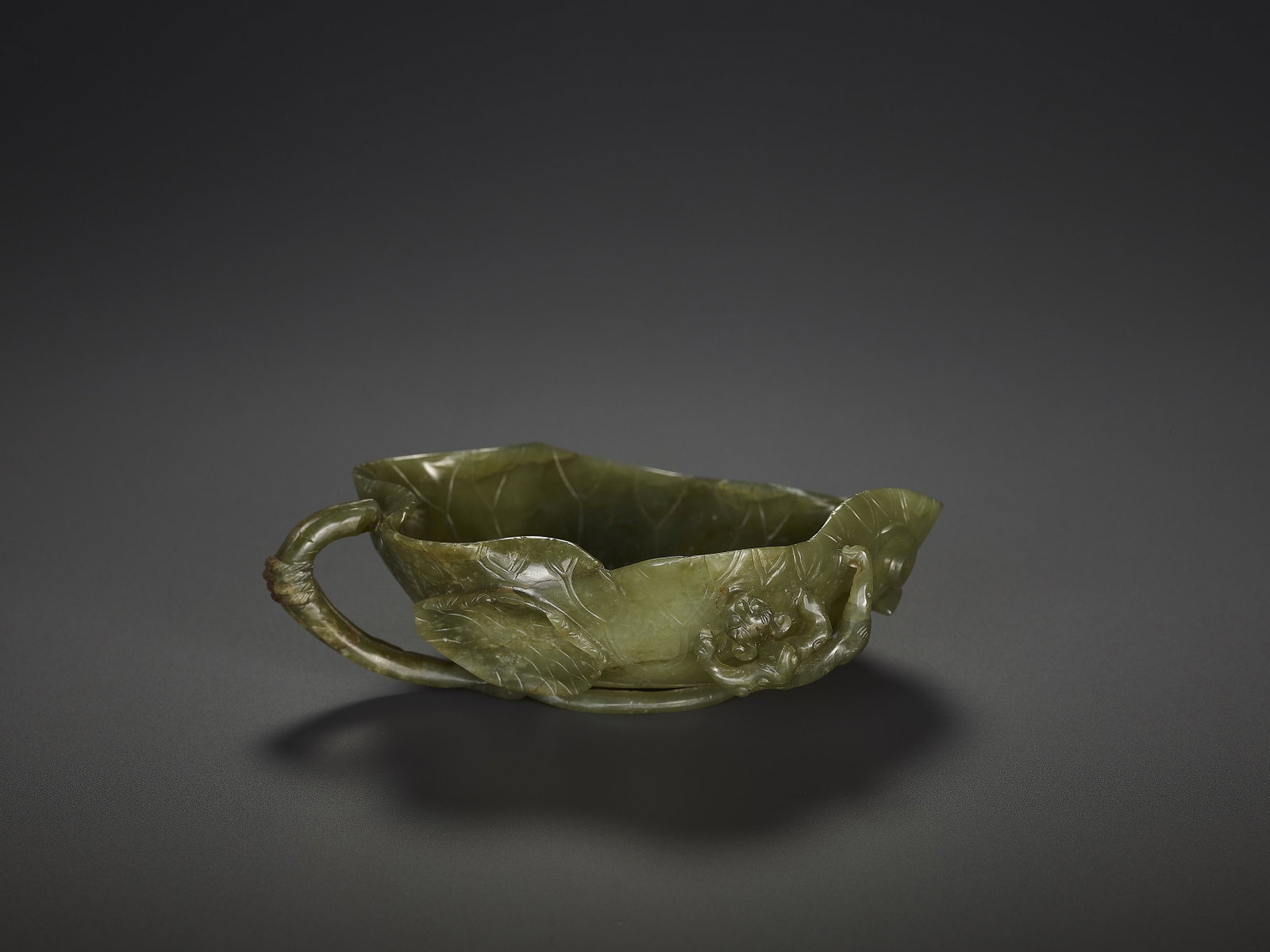 A SPINACH JADE 'LOTUS' WASHER, QING: A SPINACH JADE 'LOTUS' WASHER, QING China, 19th century. The translucent stone of dark green color with a few dispersed grey inclusions. Carved in openwork as a large lotus leaf sided by a smaller lea