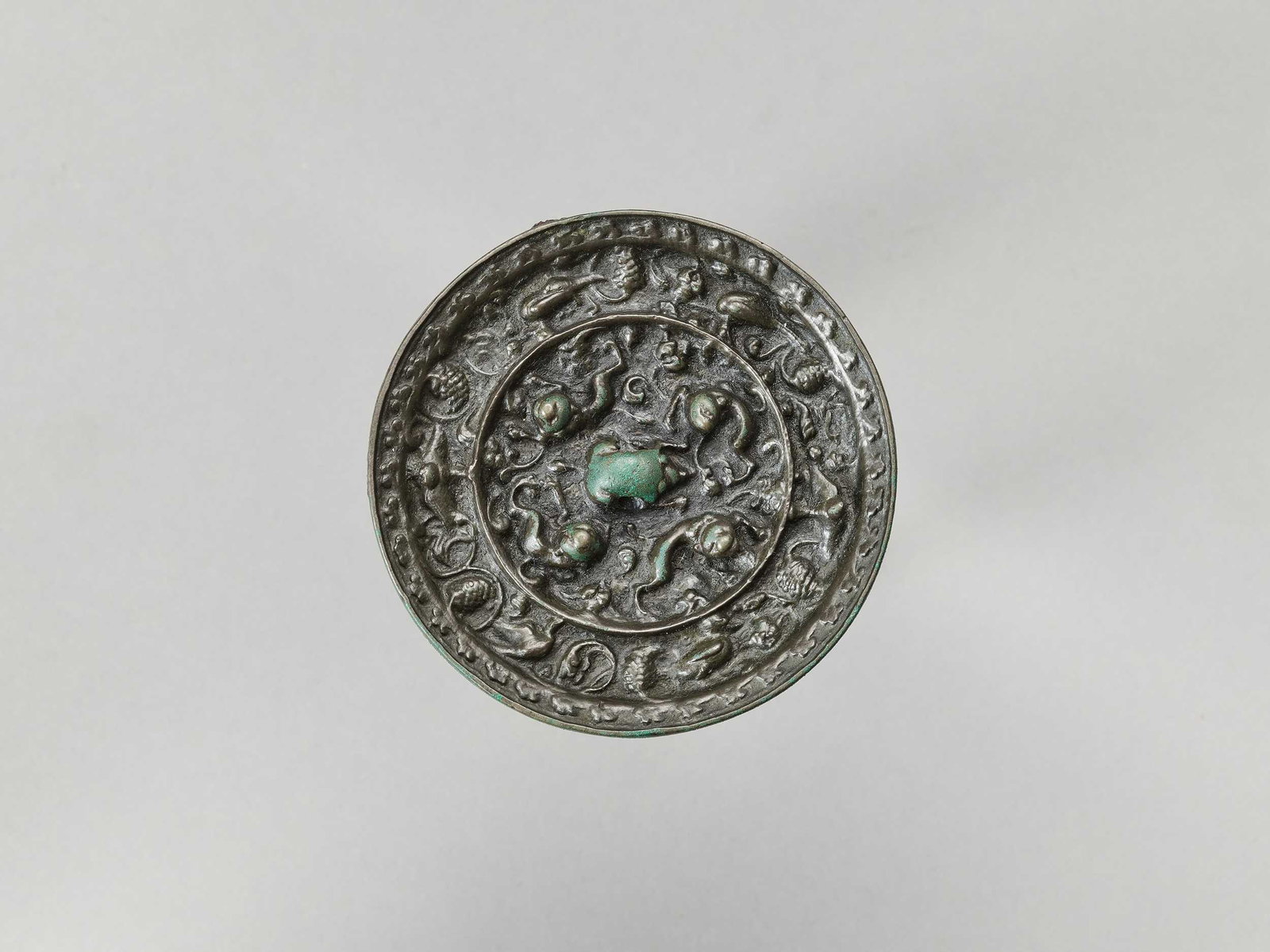 A BRONZE 'LION AND GRAPEVINE' MIRROR, FIVE DYNASTIES: A BRONZE 'LION AND GRAPEVINE' MIRROR, FIVE DYNASTIES China, 907-979. Of circular form, the back cast in high relief in the center with a crouching beast-form loop surrounded by four lions amid grapevi
