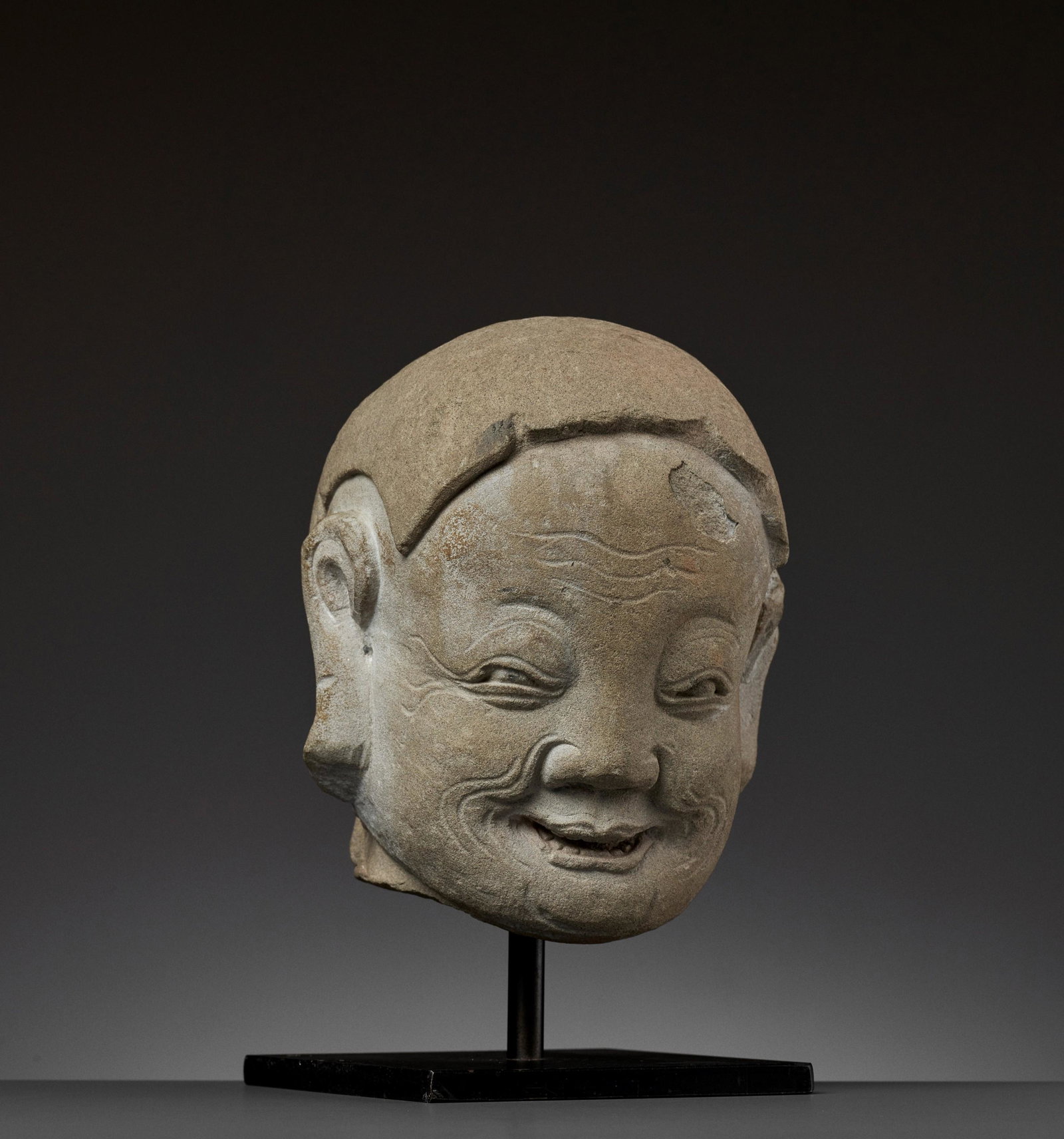 A SANDSTONE HEAD OF A LUOHAN, MING DYNASTY: A SANDSTONE HEAD OF A LUOHAN, MING DYNASTY China, 14th-15th century. Expressively carved with a broad smile and deep-cut lines, beneath a short broad nose and narrow squinting eyes, all between large 