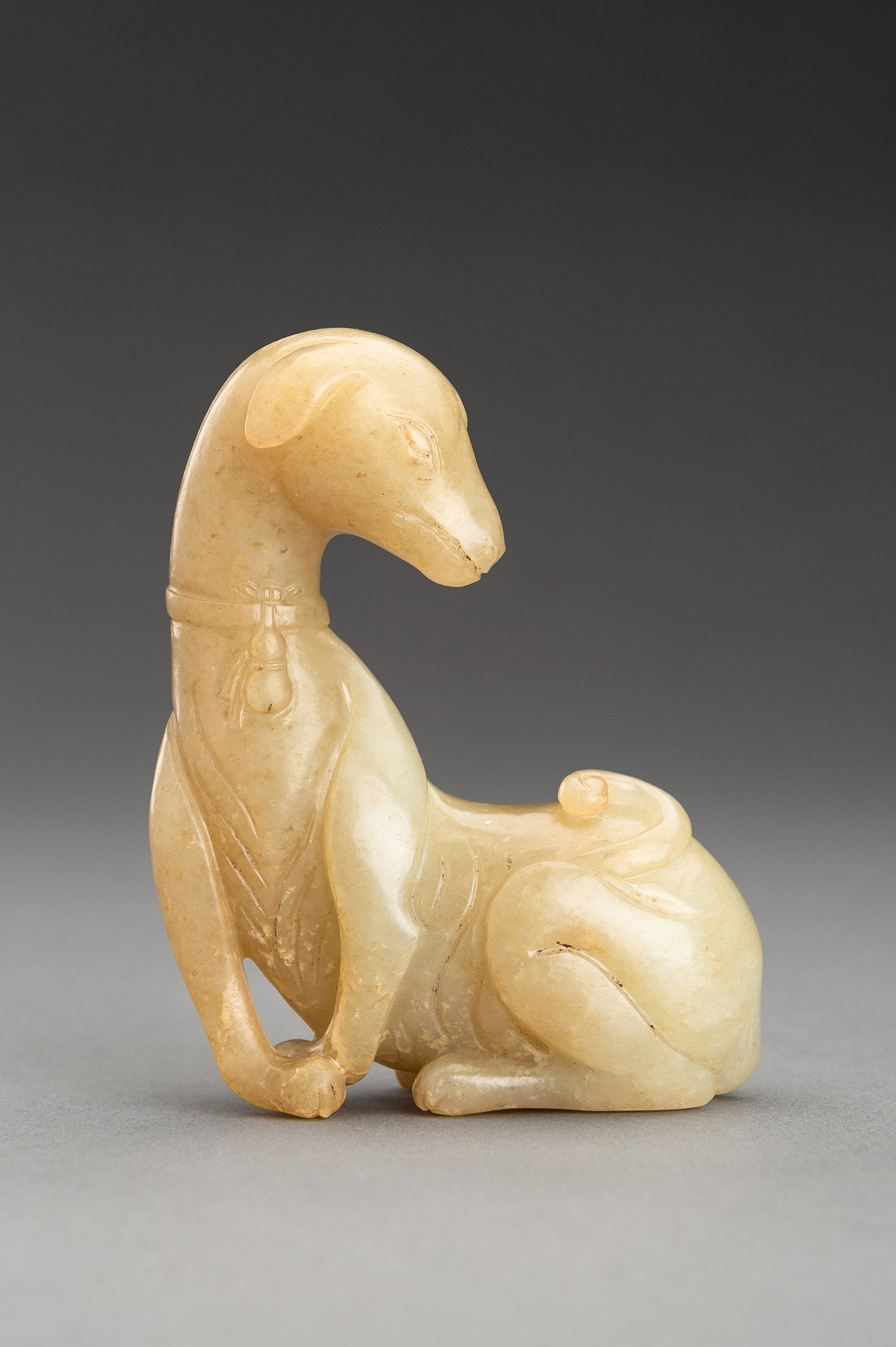 A PALE YELLOW JADE FIGURE OF A DOG, QING: A PALE YELLOW JADE FIGURE OF A DOG, QING China, Qing Dynasty (1644-1912). Elegantly seated with its front paws crossing, the head turned backwards, wearing a collar suspending a double gourd, its tail