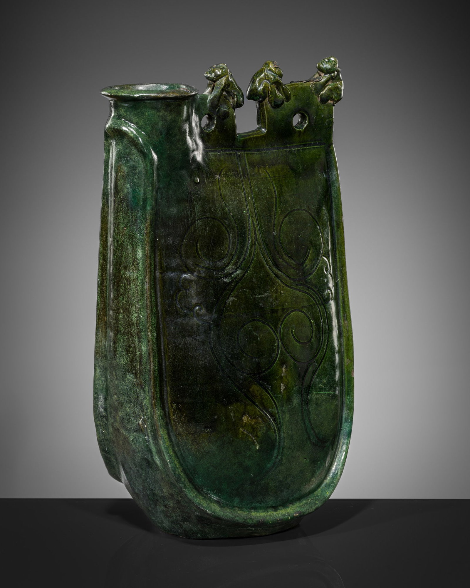 A RARE GREEN-GLAZED POTTERY FLASK, LIAO DYNASTY: A RARE GREEN-GLAZED POTTERY FLASK, LIAO DYNASTY China, 907-1125. The flask in the form of leather water or wine skin, with stiff seamed edges. The sides incised with scrolling vines and the top flap n