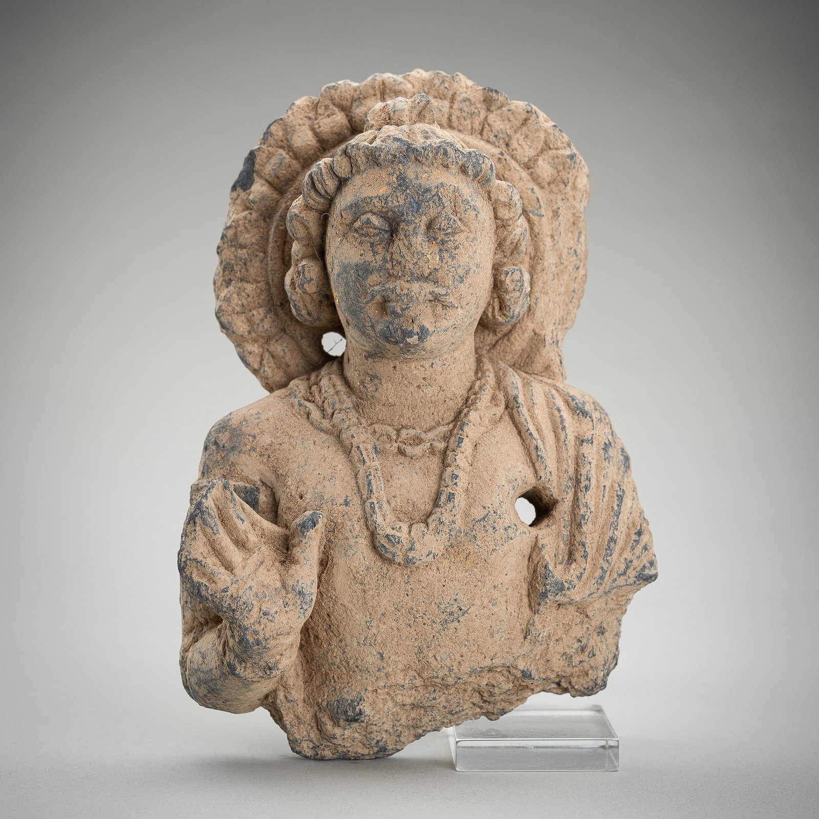 A SCHIST BUST OF A DEITY, GANDHARA, 1ST - 5TH CENTURY AD: A SCHIST BUST OF A DEITY, GANDHARA, 1ST - 5TH CENTURY AD Ancient region of Gandhara. Depicted with a serene expression, one hand raised in abhaya mudra, the stout body adorned with beaded jewelry and 