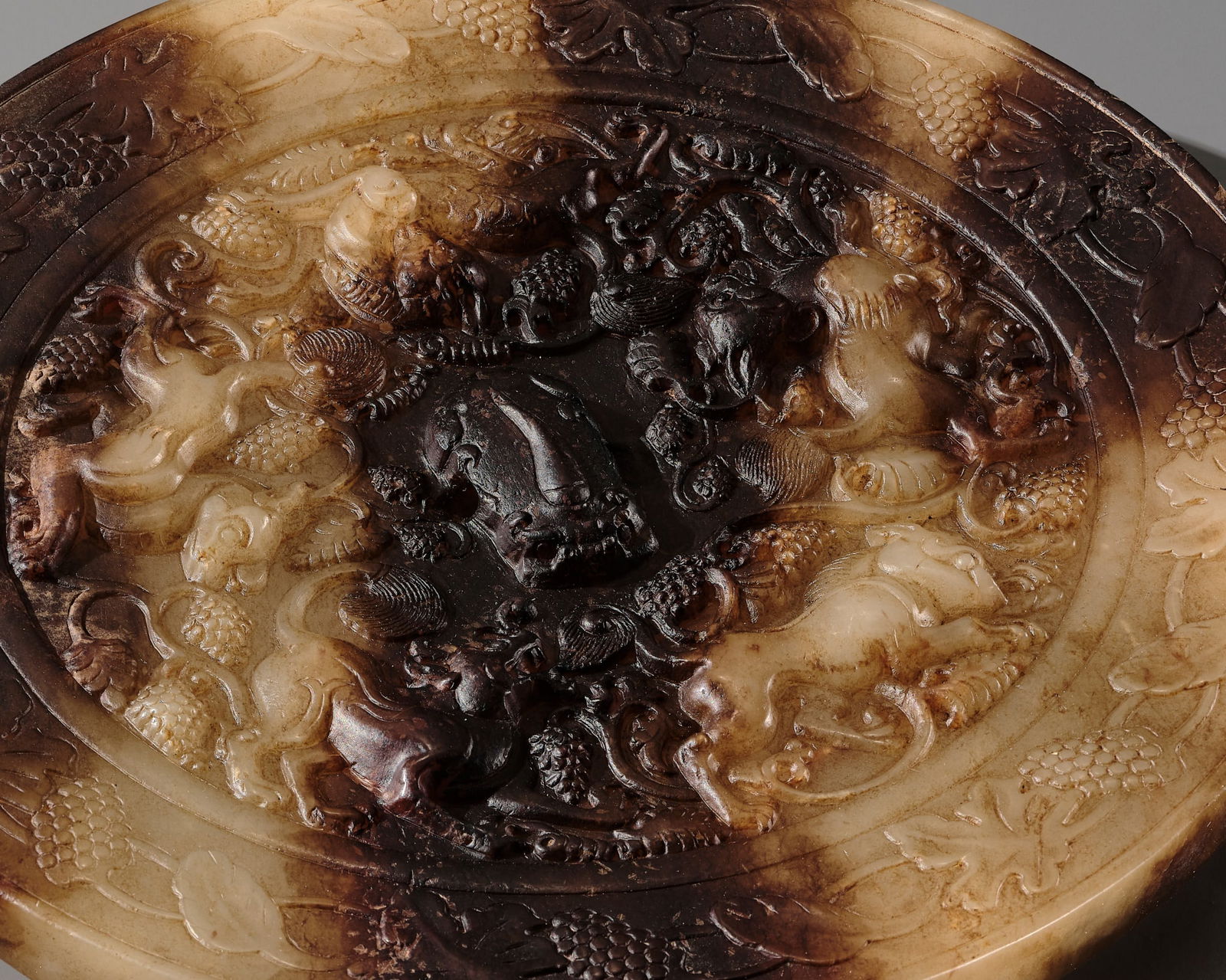 A VERY RARE YELLOW AND BROWN JADE 'LION AND GRAPEVINE' MIRROR, QING DYNASTY OR EARLIER: A VERY RARE YELLOW AND BROWN JADE 'LION AND GRAPEVINE' MIRROR, QING DYNASTY OR EARLIER China. Finely carved, of circular form with a gently concave section, the backside centered by a crouching beast