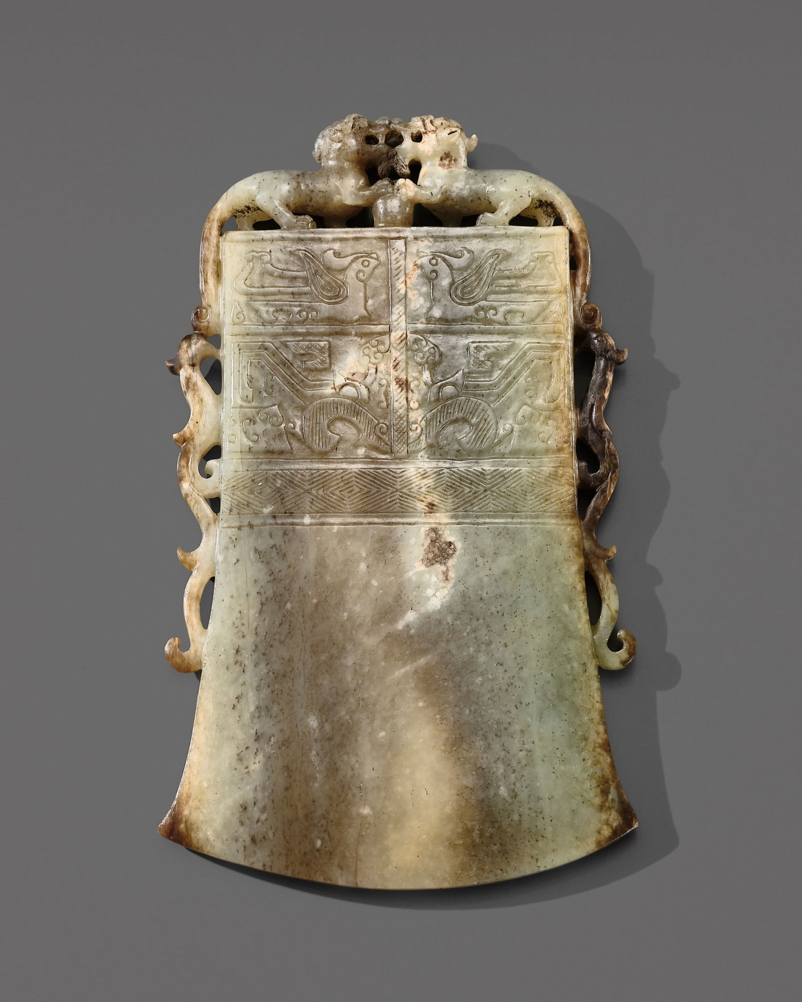 AN ARCHAISTIC CELADON AND BROWN JADE 'AXE-HEAD' ORNAMENT, QING DYNASTY: AN ARCHAISTIC CELADON AND BROWN JADE 'AXE-HEAD' ORNAMENT, QING DYNASTY China, 18th century. Finely carved as a bell-shaped axe head, its surface neatly detailed on both sides with symmetrical registe