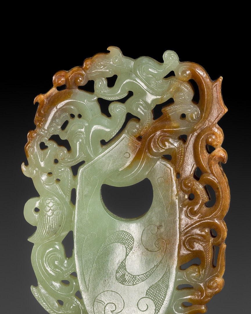 AN OPENWORK PALE CELADON AND RUSSET 'ARCHER'S RING AND BEASTS' PENDANT, HAN DYNASTY: AN OPENWORK PALE CELADON AND RUSSET 'ARCHER'S RING AND BEASTS' PENDANT, HAN DYNASTY China, 206 BC-220 AD. Finely carved and pierced, the domed plaque centers on an archer's-ring-shaped shield encircl