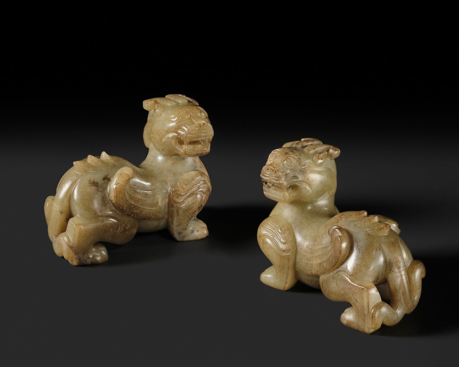 A PAIR OF PALE CELADON AND BROWN JADE 'BIXIE' FIGURES, HAN DYNASTY: A PAIR OF PALE CELADON AND BROWN JADE 'BIXIE' FIGURES, HAN DYNASTY China, 206 BC-220 AD. Carved in a mirror image, each beast with raised head, open mouth revealing teeth and tongue, a pair of curvin