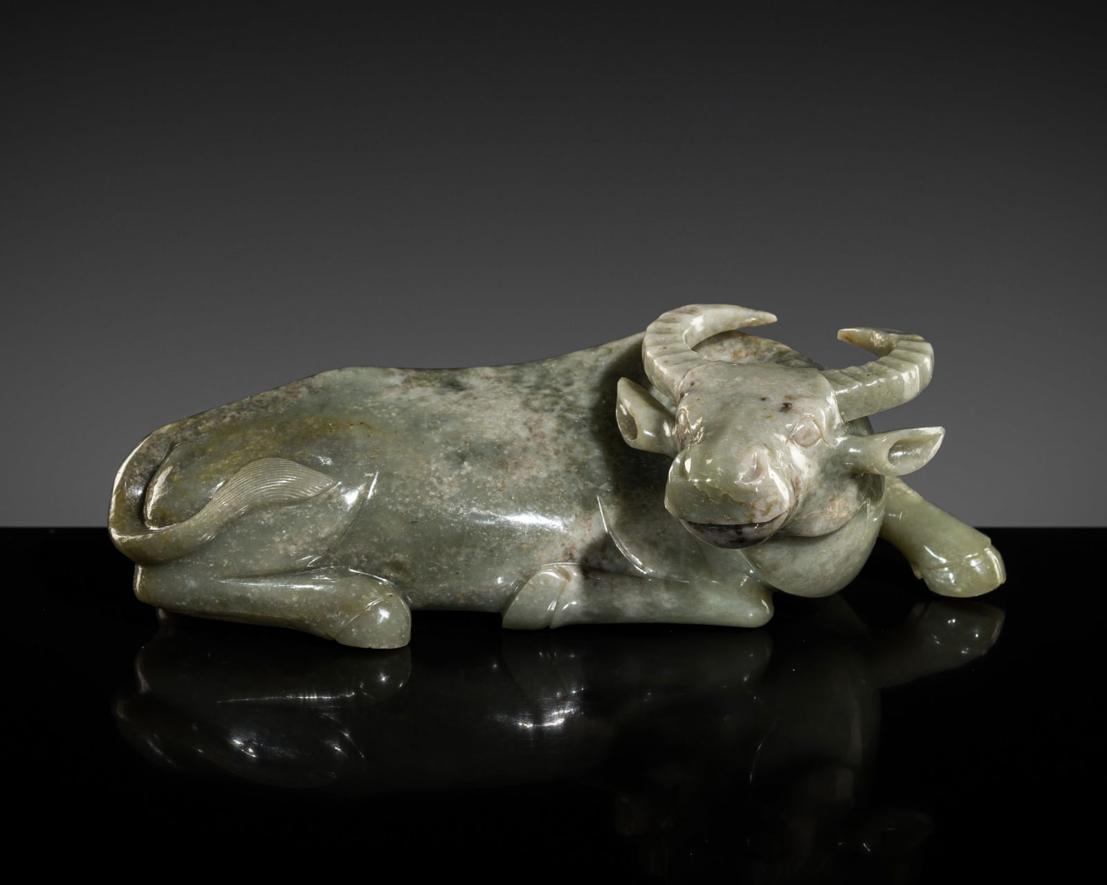 A GRAYISH-GREEN JADE FIGURE OF A WATER BUFFALO, LATE QING DYNASTY-REPUBLIC PERIOD: A GRAYISH-GREEN JADE FIGURE OF A WATER BUFFALO, LATE QING DYNASTY-REPUBLIC PERIOD China, circa 1850-1950. Powerfully carved in a recumbent pose, with the right foreleg extended and the other tucked un