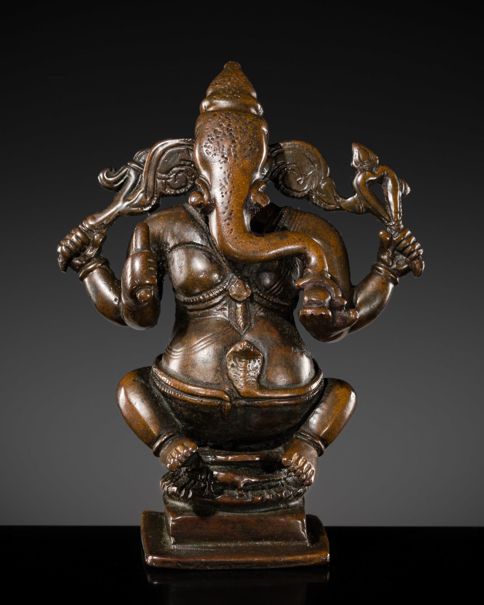 A BRONZE FIGURE OF GANESHA, SOUTH INDIA, 17TH-18TH CENTURY: A BRONZE FIGURE OF GANESHA, SOUTH INDIA, 17TH-18TH CENTURY The potbellied deity seated on a waisted lotus base raised over a waisted pedestal, his four arms radiating around him holding a goad, broken