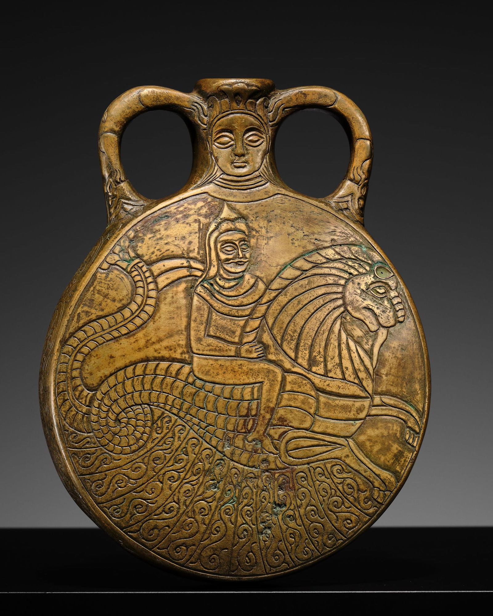 A FINELY ENGRAVED BRASS 'HERO ON MYTHICAL BEAST' PILGRIM FLASK, SASANIAN EMPIRE: A FINELY ENGRAVED BRASS 'HERO ON MYTHICAL BEAST' PILGRIM FLASK, SASANIAN EMPIRE Iran, 224-651 AD. Well cast with a flattened circular body surmounted by a short cylindrical neck set with a pair of bea