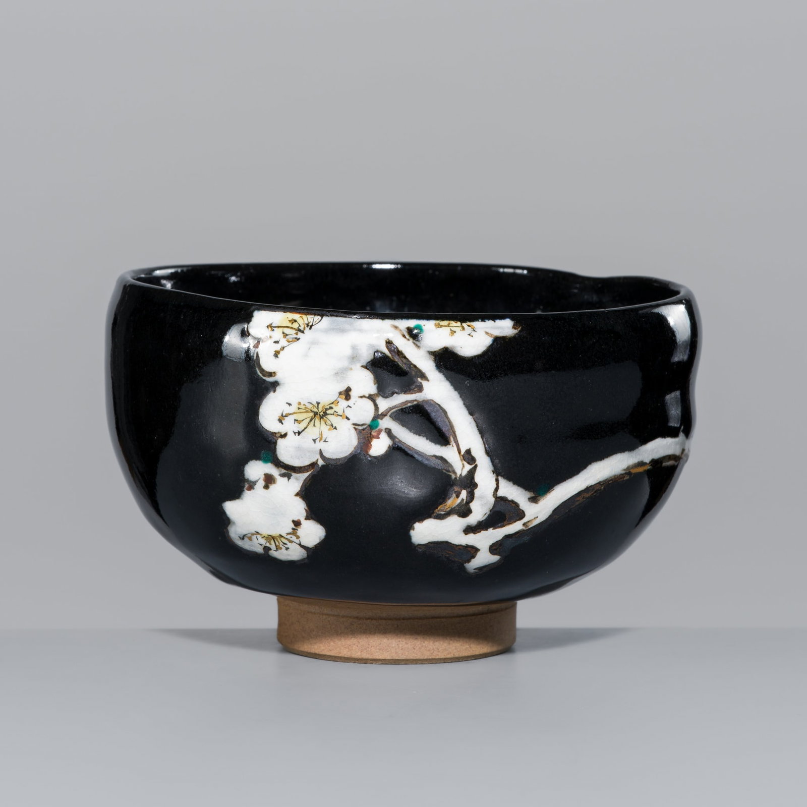 MAKUZU KOZAN I: A BLACK GLAZED STUDIO PORCELAIN 'SNOWY PLUM' CHAWAN (TEA BOWL): MAKUZU KOZAN I: A BLACK GLAZED STUDIO PORCELAIN 'SNOWY PLUM' CHAWAN (TEA BOWL) By Makuzu Kozan I (1842-1916), signed Kozan and Makuzu Japan, c. 1916 The bowl of irregular form with an indentation to s