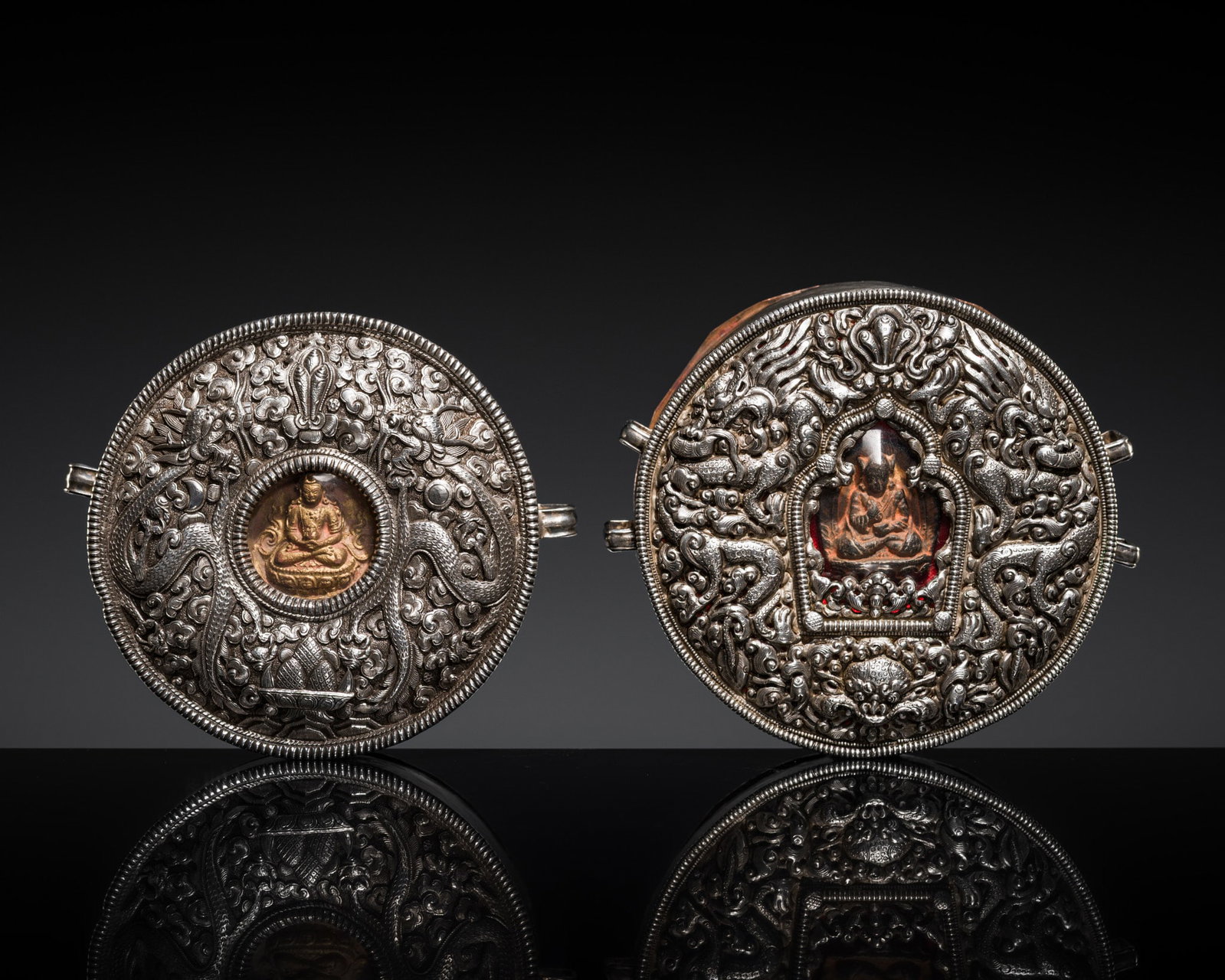 TWO FINE EMBOSSED SILVER AND COPPER PORTABLE SHRINES WITH TSATSA, GAU: TWO FINE EMBOSSED SILVER AND COPPER PORTABLE SHRINES WITH TSATSA, GAU Tibet, 19th century. Each of circular form with a domed cover framed by beaded rims, detailed to the top with a pair of sinuous dr
