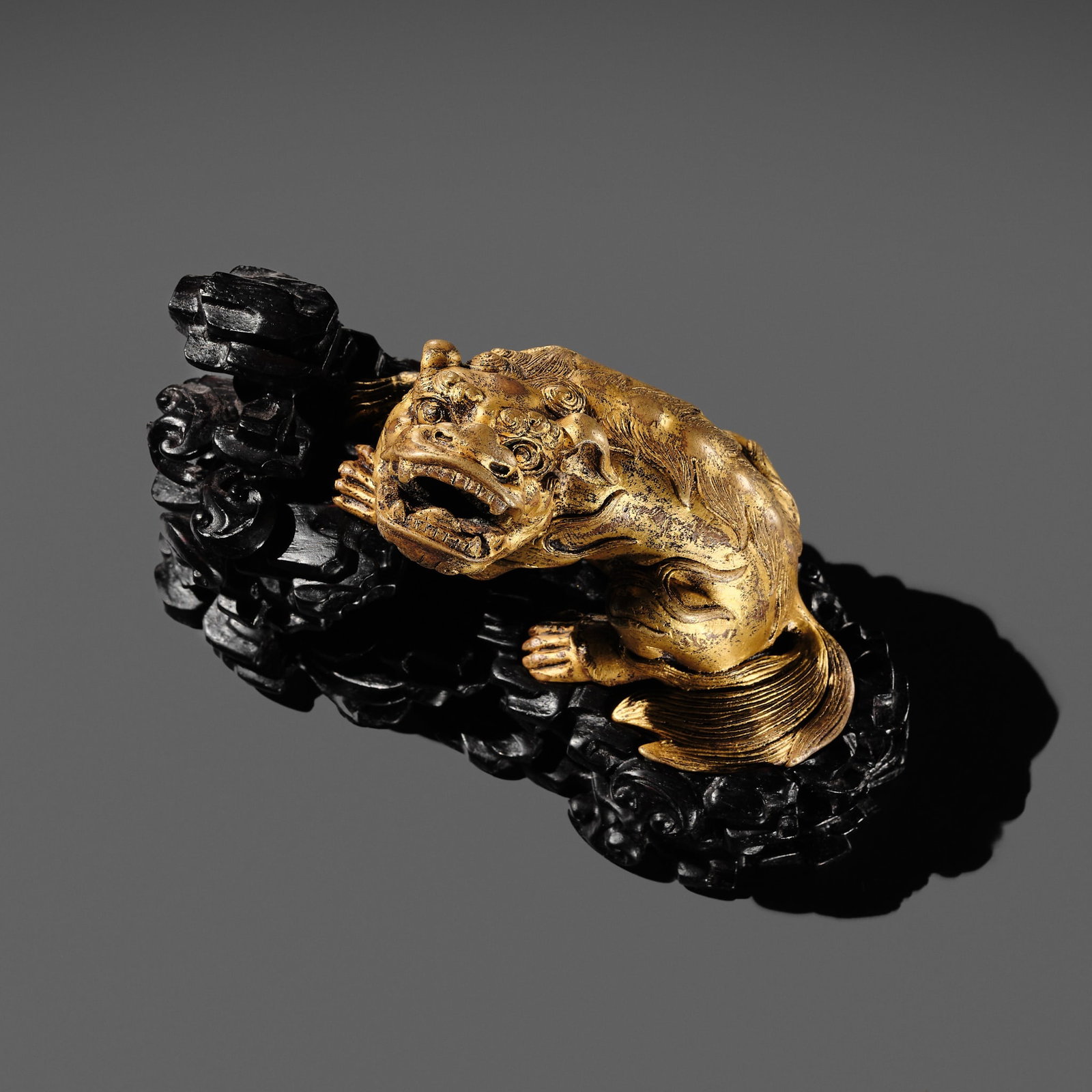 A GILT BRONZE 'LION' WEIGHT WITH A MATCHING ZITAN BASE, CHINA, 16TH-17TH CENTURY: † A GILT BRONZE 'LION' WEIGHT WITH A MATCHING ZITAN BASE, CHINA, 16TH-17TH CENTURY Finely cast in a crouching position, its face turned to the left, the mouth ajar revealing sharp fangs, the ear