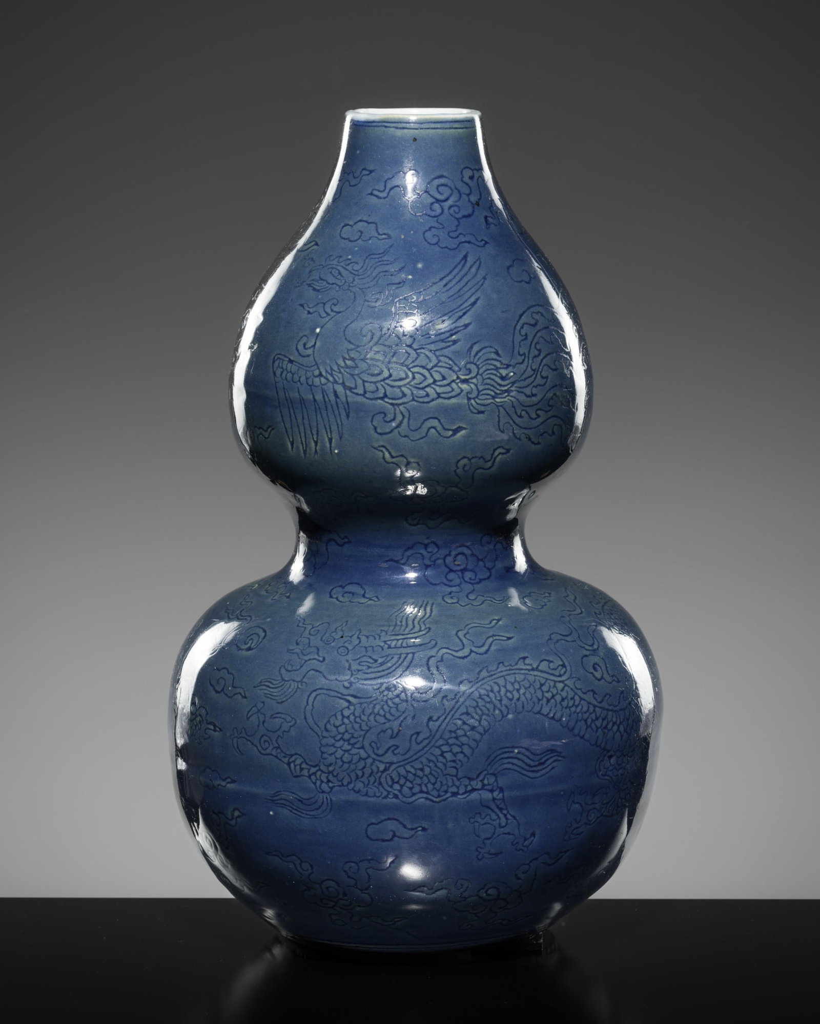 AN INCISED BLUE-GLAZED 'DRAGON AND PHOENIX' DOUBLE GOURD VASE, LATE MING DYNASTY: AN INCISED BLUE-GLAZED 'DRAGON AND PHOENIX' DOUBLE GOURD VASE, LATE MING DYNASTY China, 16th-17th century. Well potted of double gourd form, rising from a flat circular foot to a tall neck, the lower 