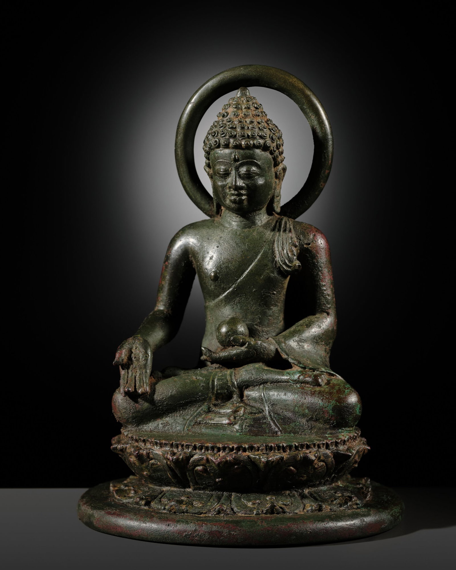 A BRONZE FIGURE OF THE MEDICINE BUDDHA (BHAISAJYAGURU), CENTRAL JAVANESE PERIOD, 9TH-10TH CENTURY: A BRONZE FIGURE OF THE MEDICINE BUDDHA (BHAISAJYAGURU), CENTRAL JAVANESE PERIOD, 9TH-10TH CENTURY Indonesia. Seated in dhyanasana on a double-lotus base raised on a wide cushion, his right hand lowere