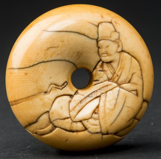 MANJUGATA WITH SCHOLAR: Netsuke, ivory. Japan, 19th century The upwards facing side of this Manjugata netsuke model in the style of the Osaka school (like Naomitsu), shows a scholar seated on a cushion, carved in relief. On 